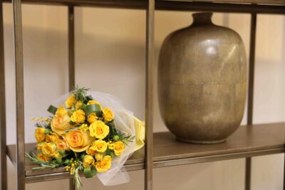 Buquê de rosas amarelas encima de uma estante ao lado de vaso marrom.